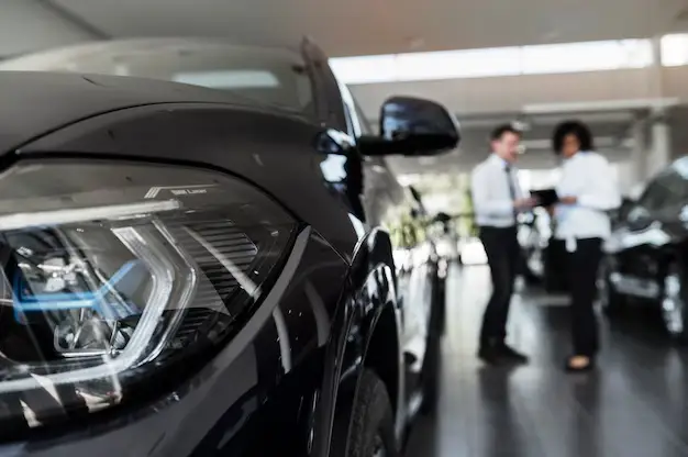 A shiny black car’s headlamp close up while A man and a woman discuss something on a book in the background