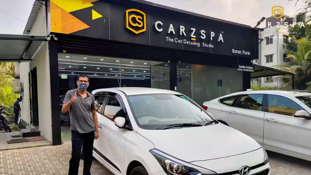 A man stands near a white sedan in front of a car detailing studio while showing a thumbs-up sign of approval