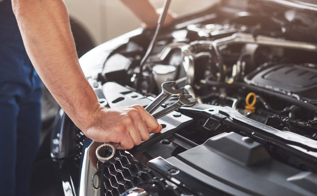 Image alt text: the open bonnet of car with tools lying on and the muscular arm of a worker is visible in the frame